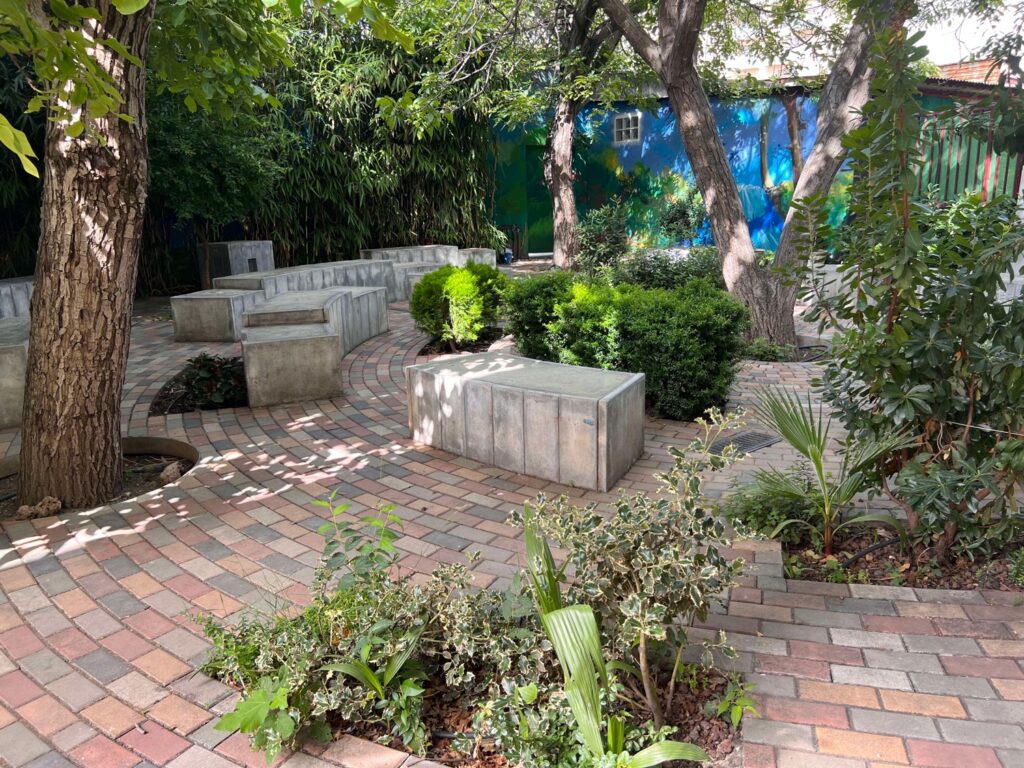 Área verde con bancos de concreto y caminos de adoquines, rodeada de árboles y arbustos.