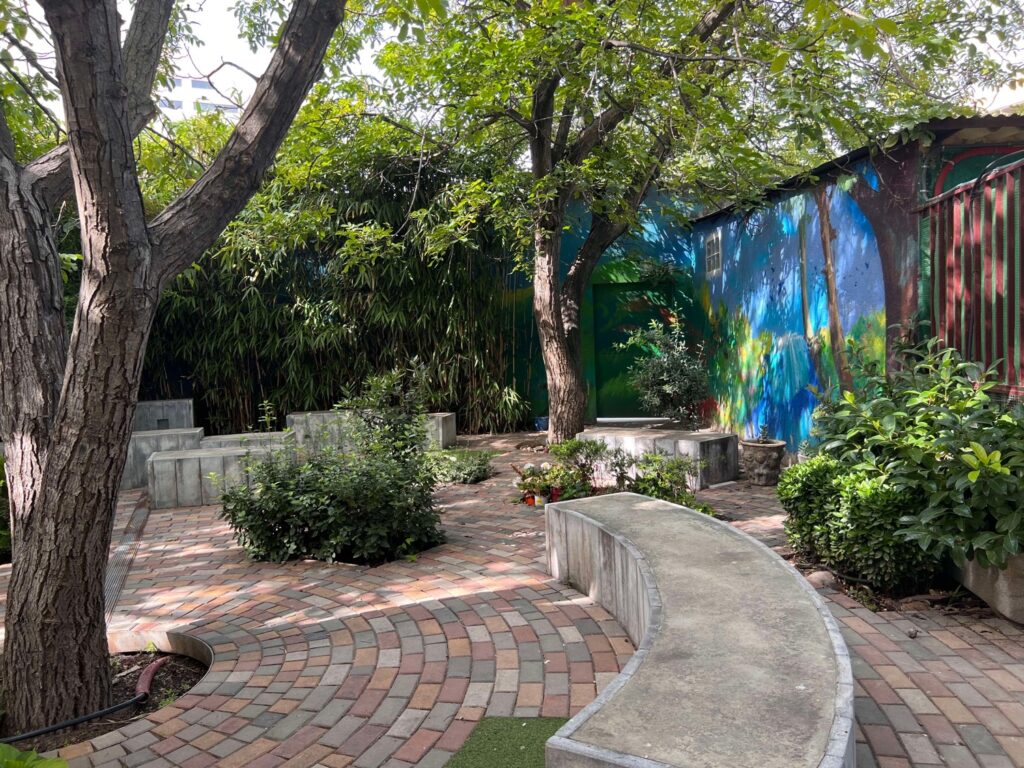 Jardín con adoquines, bancos de cemento, árboles frondosos y mural colorido en la pared. Ambiente tranquilo y sombreado.