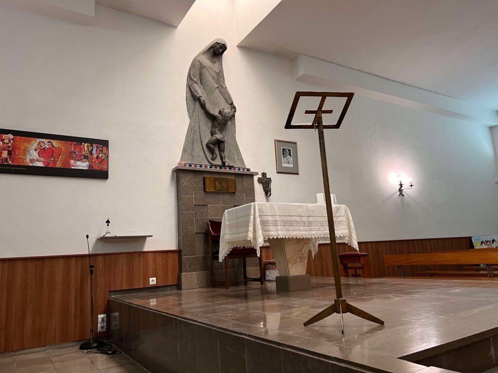 Interior de una iglesia con altar, estatua religiosa, soporte de micrófono y cuadro en la pared. Iluminación cálida.