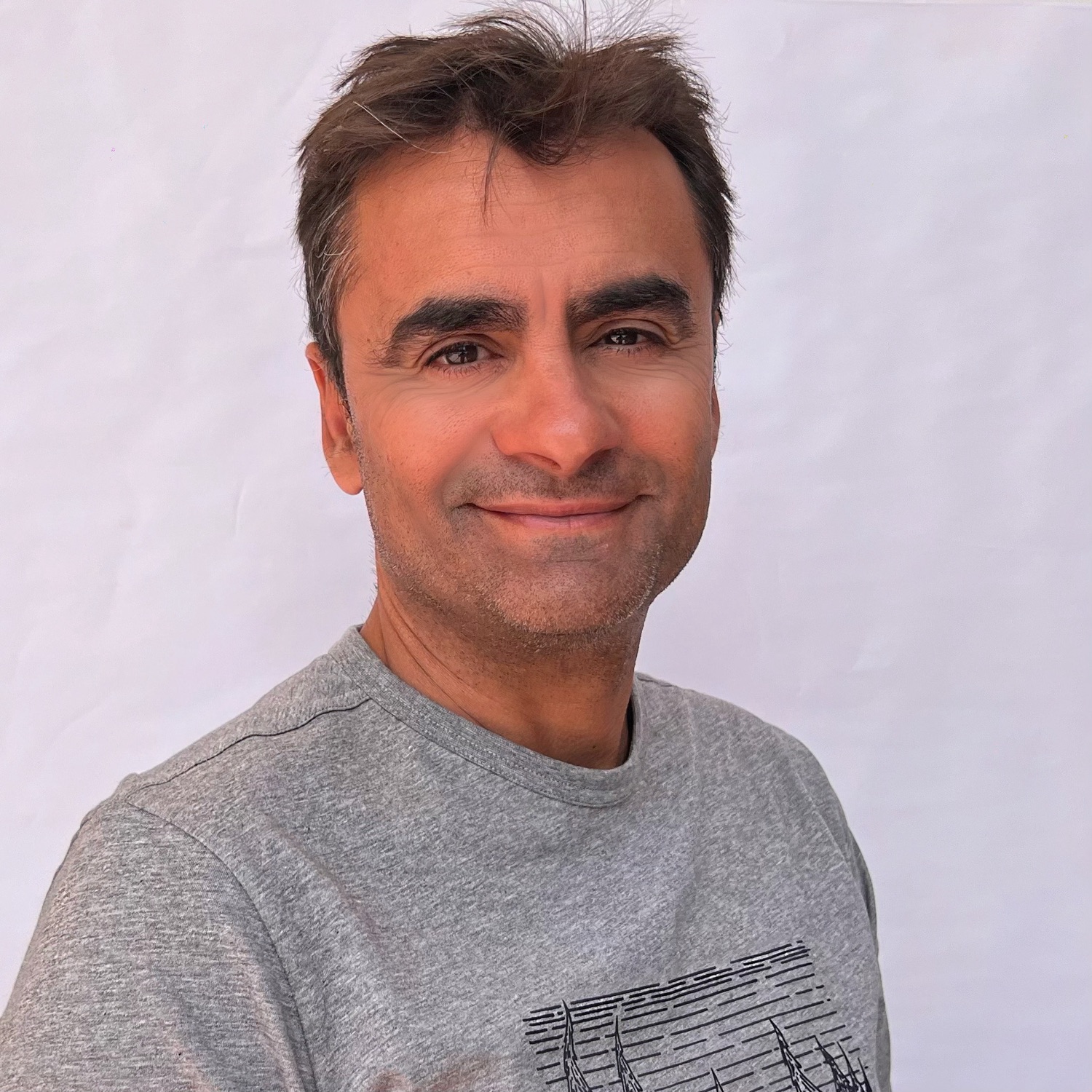 Hombre de cabello oscuro con camiseta gris, sonriendo frente a un fondo blanco.