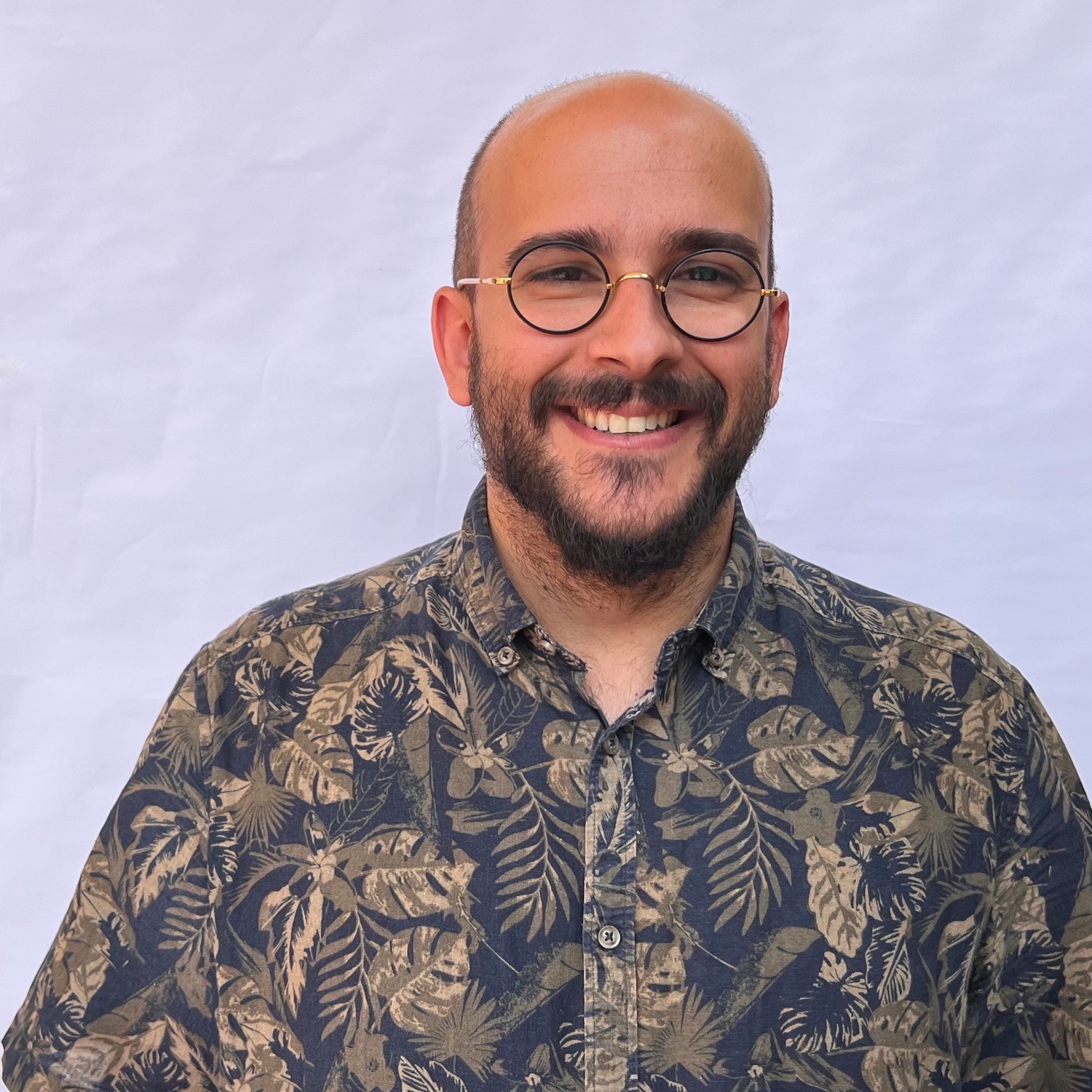 Hombre sonriendo con camisa estampada tropical y gafas redondas, fondo blanco liso.