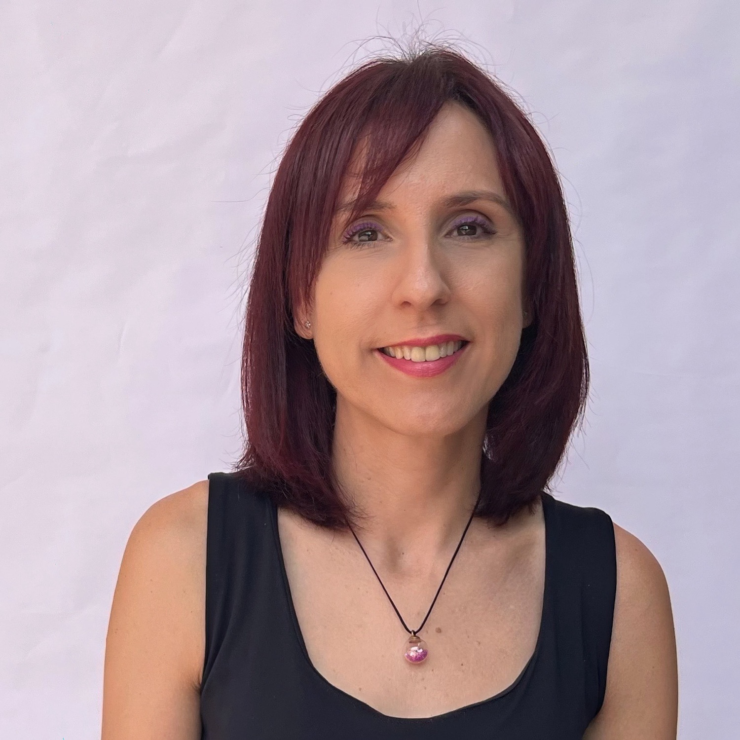 Mujer sonriendo con cabello castaño rojizo, viste blusa negra y collar, sobre fondo blanco liso.