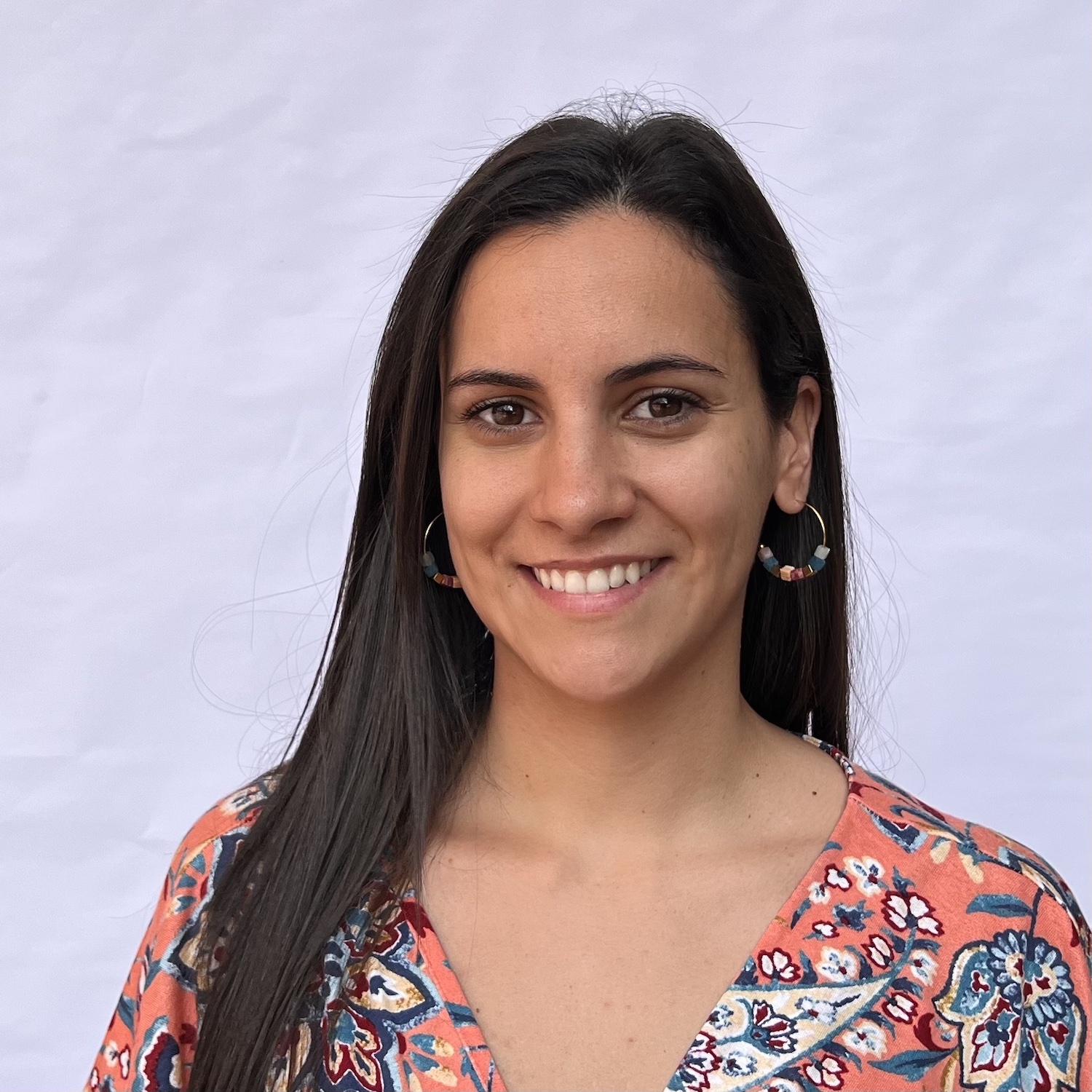 Mujer con cabello largo y lacio, sonríe al cámara, con camisa floral naranja y aretes de aro. Fondo blanco.