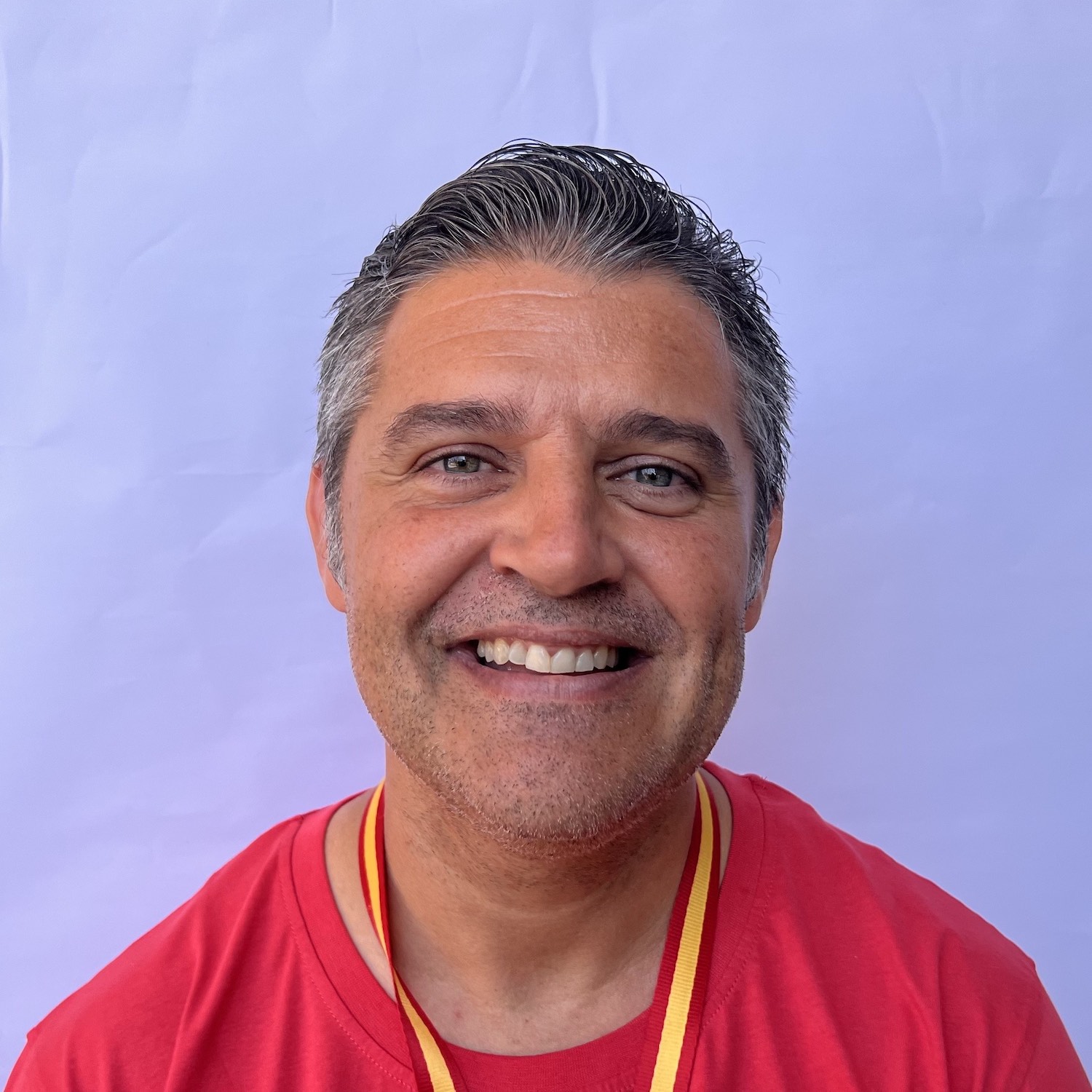 Hombre sonriente con camiseta roja y medalla, frente a un fondo azul claro.