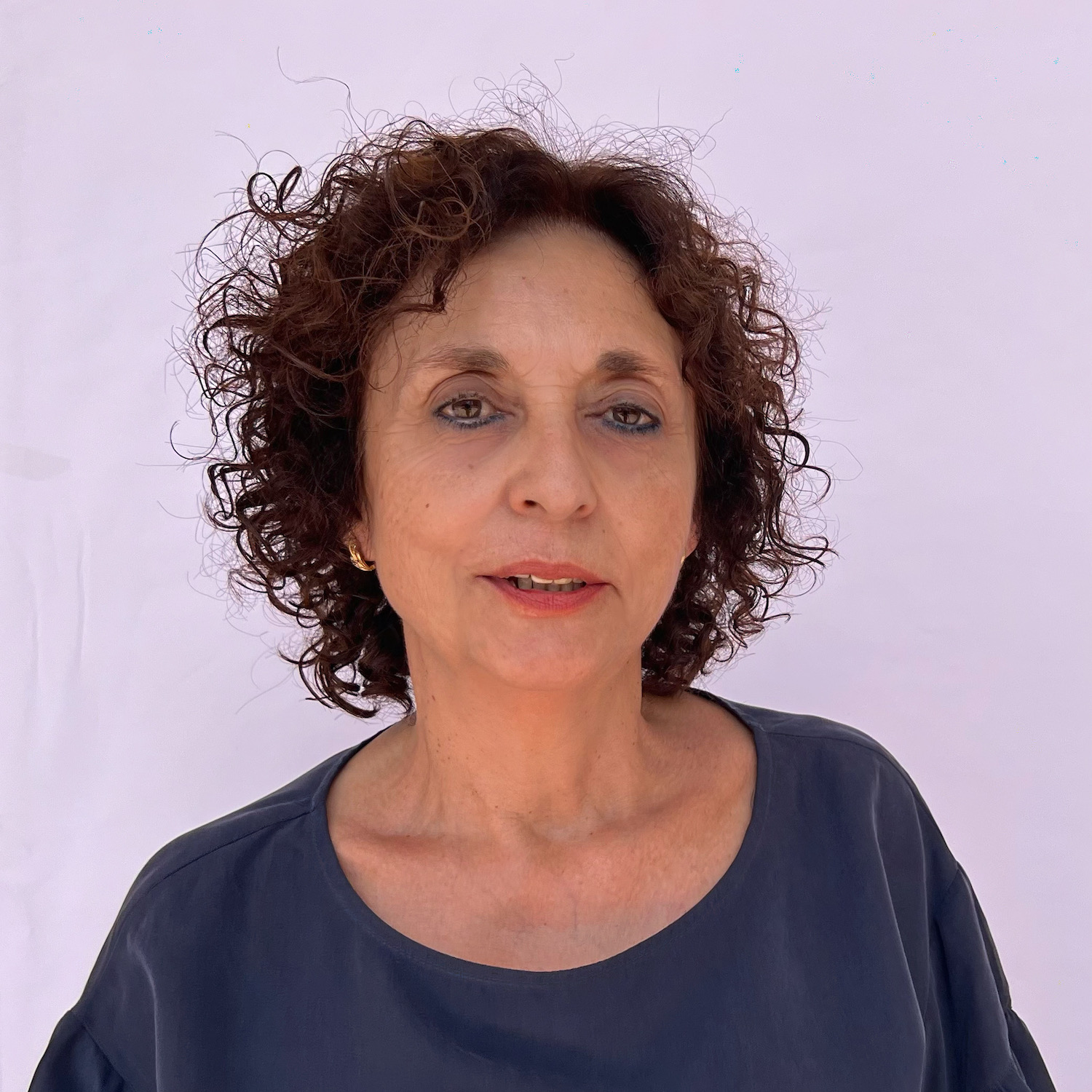 Mujer de cabello rizado y corto con blusa oscura, posa frente a un fondo claro.
