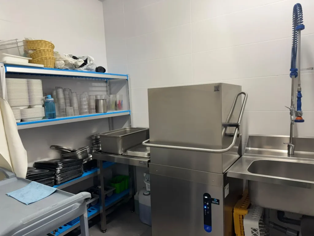 Vista general de la zona de fregado de la cocina, con lavavajillas industrial, fregadero y estanterías de menaje